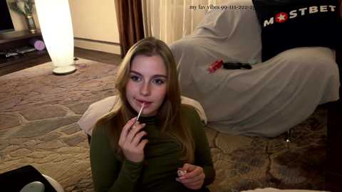 Media: Video of a young Caucasian woman with straight blonde hair, wearing a green top, licking a lollipop while seated on a patterned carpet in a dimly lit room.