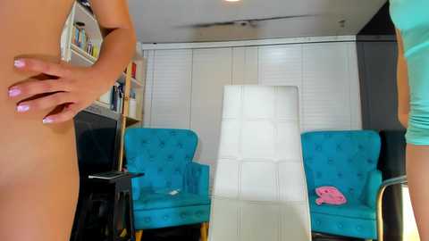 Media: Video of a nude woman with fair skin and pink nails, standing in a modern living room with turquoise armchairs, white furniture, and a white wall.