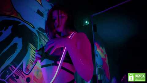 Media: Video of a curvy, light-skinned woman with long, wavy brown hair in a dark, urban alley. She wears a black bra and pink neon lights illuminate her body, with colorful graffiti and a green DMC logo in the background.