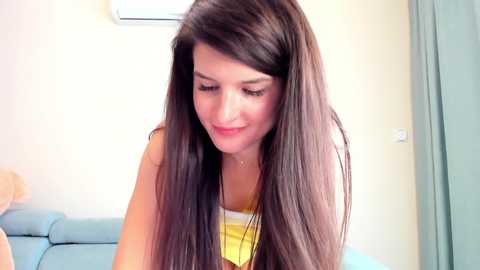 Media: Video of a young woman with long, straight brown hair, wearing a yellow tank top, smiling, sitting on a light blue couch in a modern, well-lit room.