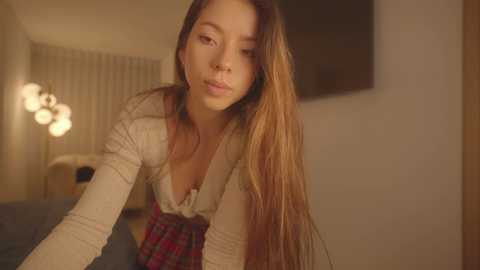 Media: Video of a young, light-skinned woman with long, straight brown hair, wearing a beige top and a plaid skirt, leaning forward in a dimly lit room with a glowing lamp and soft, muted tones.