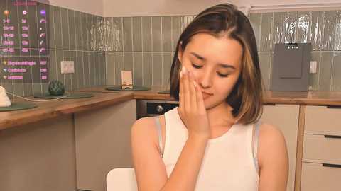 Media: Video of a young, light-skinned woman with shoulder-length brown hair, wearing a white tank top, covering her eyes with her hands in a modern kitchen with green tiles and white cabinets.