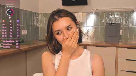 Media: Video of a young woman with fair skin and straight brown hair, wearing a white tank top, covering her mouth with her hand, in a modern kitchen with tiled backsplash.