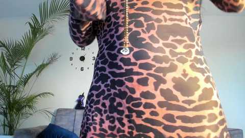 Media: A video of a person wearing a leopard print dress, standing in a modern living room with a clock wall art, palm tree, and a cat on a sofa.