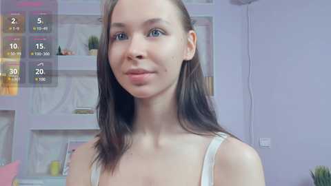 Media: Video of a young Caucasian woman with light skin, straight brown hair, wearing a white tank top, standing indoors with a lavender wall and bookshelves in the background.