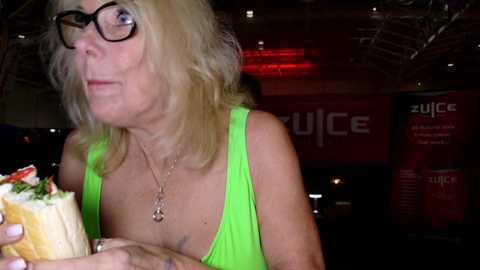 Media: Video of a middle-aged woman with shoulder-length blonde hair, wearing glasses and a bright green tank top, eating a sandwich. Background shows a dimly lit room with red lighting and blurred text.