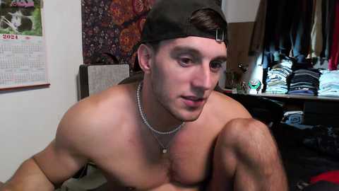 Media: Video of a shirtless young man with short dark hair, wearing a backward baseball cap and silver necklace, leaning on a bed with a calendar and a colorful tapestry on the wall.