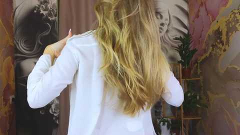 Media: Video of a woman with long, wavy blonde hair, wearing a white long-sleeved shirt, standing in a room with maroon and gray walls featuring abstract art and a green plant on a wooden shelf.