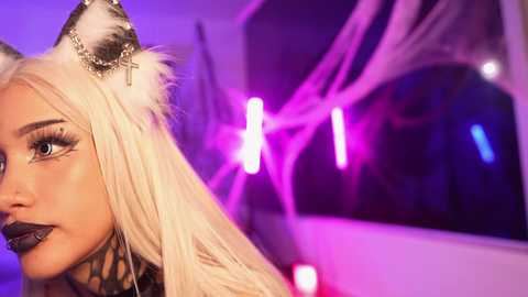 Media: Video of a young woman with long platinum blonde hair, wearing cat ears and dramatic black makeup, under neon purple and pink lights.