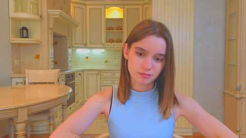 Media: Video of a young woman with straight brown hair, wearing a light blue sleeveless top, standing in a cozy, vintage-style kitchen with wooden cabinets and a round table.