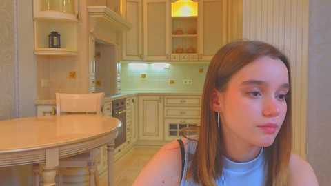 Media: Video of a young girl with straight brown hair, wearing a light blue top, standing in a modern, cream-colored kitchen with wooden cabinetry, round table, and pendant light.