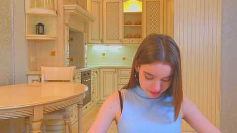 Media: A video of a young woman with straight brown hair, wearing a light blue sleeveless top, standing in a modern, cream-colored kitchen with wooden cabinets and a round wooden dining table.