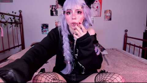 Media: Video of a pale-skinned, long-haired woman with lavender hair, wearing a black hoodie, fishnet stockings, and black gloves, sitting on a bed in a pink room with posters on the wall.