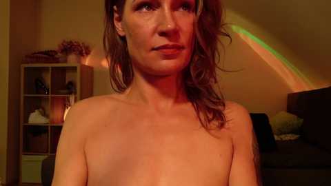 Media: Video of a topless woman with light skin, brown hair, and a thoughtful expression, standing indoors. Background features a wooden bookshelf and a bed with a green blanket. Warm, ambient lighting.