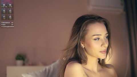 Media: Video of a young woman with long brown hair, wearing a white top, looking thoughtful. Background includes a bed with a potted plant and a digital screen displaying a live stream.