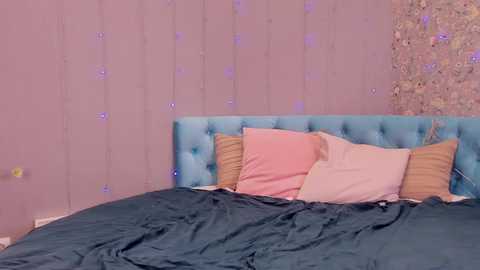 Media: Video of a bed with a teal tufted headboard, pink pillows, and a teal blanket, set against a pastel pink wall adorned with fairy lights.