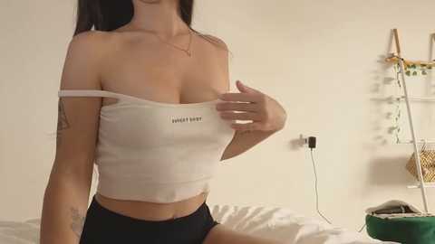 Media: Video of a light-skinned woman with long black hair, wearing a white off-shoulder crop top and black shorts, seated on a bed in a dimly lit bedroom with a green plant and wall-mounted shelves.