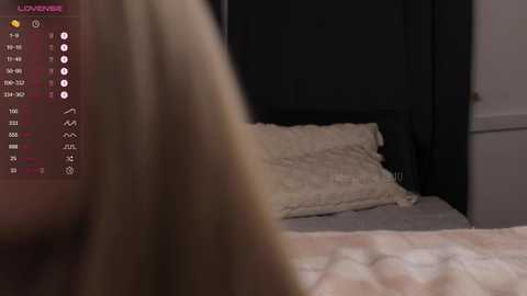 Media: A video of a dimly lit bedroom with a blonde woman in the foreground, facing away, and a bed with a white blanket and pillows in the background.