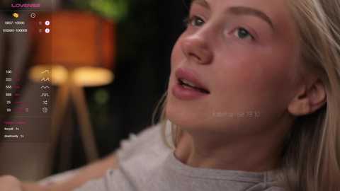 Media: A close-up video of a blonde woman with fair skin, wearing a grey top, looking up with a slight smile, in a dimly lit room with a lamp and blurred background.