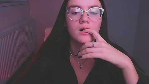 Media: Video of a young woman with fair skin, long black hair, and large glasses, biting her finger, wearing a black dress, necklace, and rings, in a dimly lit room with a radiator.