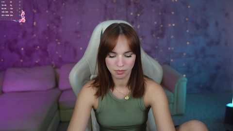 Media: Video of a young woman with medium-length brown hair, fair skin, and a slender build, wearing a green tank top, sitting on a white gaming chair in a dimly lit room with purple and blue accents.