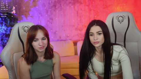 Media: Video of two young women in gaming chairs with a colorful, abstract wall backdrop. One has long red hair, wearing a green top; the other has long black hair, wearing a white top.