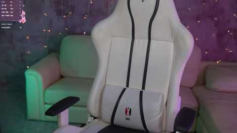 Media: Video of a white gaming chair with black armrests and a \"Gamer\" logo, set in a dimly lit room with purple and green lighting, featuring a beige sofa and a dark, textured wall adorned with fairy lights.