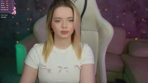 Media: A video of a young blonde woman with light skin wearing a white T-shirt with pink bows. She sits in a white gaming chair in a dimly lit room with purple and green lights.