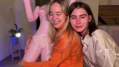 Media: Video of two women, one in a pink latex dress with polka dots, and the other in an orange latex bodysuit, smiling and embracing indoors with a plant and kitchen background.