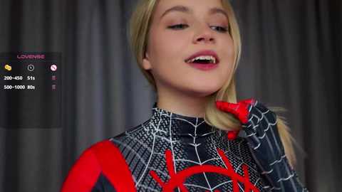 Media: Video of a young woman in a Spider-Man costume, with blonde hair, red lips, and a playful expression, against a dark curtain background.