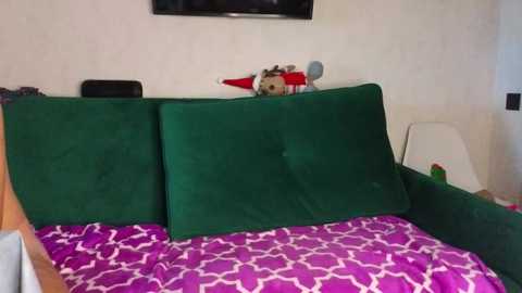 Media: Video of a green velvet sofa with three large cushions, a purple patterned blanket, and a stuffed toy elf on a white wall.