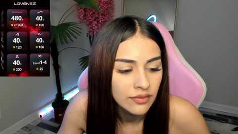 Media: Video of a young woman with long dark hair, closed eyes, sitting in a pink gaming chair. Background features a plant and digital screen displaying statistics.