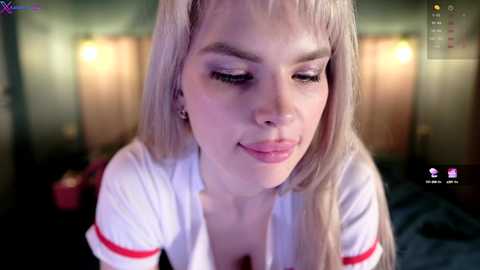 Media: Video of a young woman with light skin and long, straight blonde hair, wearing a white short-sleeve shirt with red trim. She has a slight smile and is indoors with dim lighting and blurred background.