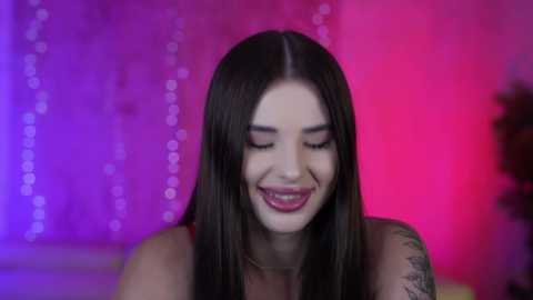 Media: Video of a smiling woman with long dark hair, light skin, and tattoos on her arm, set against a purple and pink background with string lights.