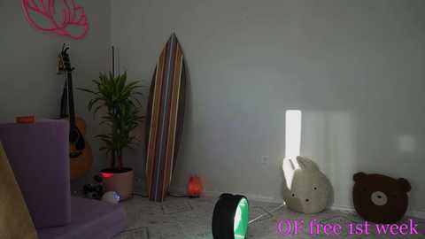 Media: Video of a dimly lit room with a purple couch, a guitar, a potted plant, a surfboard, and a stuffed bear on a gray tiled floor, with \"FREE 1st week\" text in the bottom right corner.