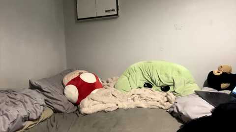 Media: A video of a cluttered bedroom with a plush green alien toy on a bed, surrounded by disheveled sheets and a stuffed bear, against a plain white wall.