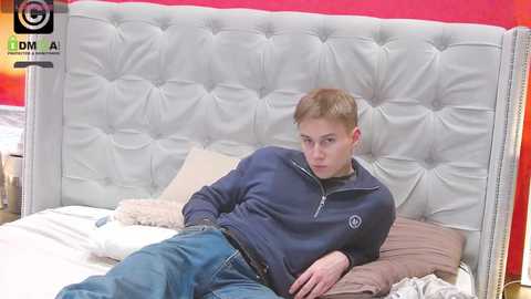 Media: Video of a young, light-skinned man with short blond hair lying on a tufted white leather headboard bed, wearing a navy zip-up hoodie and blue jeans.