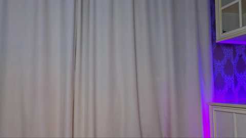 Media: Video of a room with a white curtain covering a window, a purple light illuminating the corner, and a white wooden cabinet visible on the right.