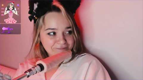 Media: Video of a smiling young woman with light brown hair and black cat ears, holding a microphone, in a dimly lit room with pink lighting.