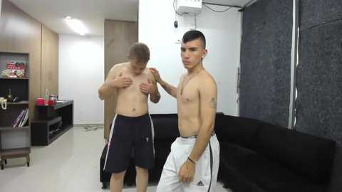 Media: Video of two shirtless men in a gym, one with short hair and dark shorts, the other with short dark hair and white shorts, both looking serious.