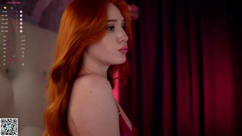 Media: Video of a fair-skinned woman with long, fiery red hair, wearing a red strapless dress, in a dimly lit room with dark curtains, focused on her profile.