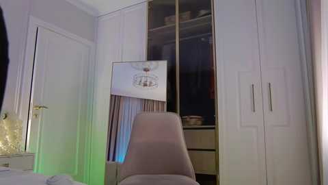 Media: Video of a modern, minimalist bedroom with white cabinets, a large mirror, and a beige chair. A framed art piece hangs on the wall, and a greenish-blue light illuminates the space.