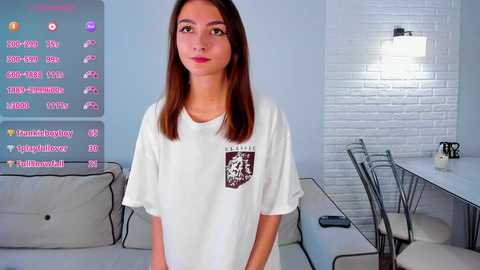 Media: Video of a young woman with long brown hair, fair skin, and wearing a white T-shirt, standing in a modern, minimalist living room with a white brick wall, glass dining table, and a digital screen displaying social media stats.