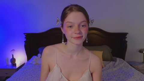 Media: Video of a young Caucasian woman with fair skin, brown hair, and a slender physique, wearing a sheer, white camisole. She sits on a bed with a dark wooden headboard and patterned bedding.