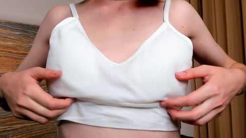 Media: Video of a fair-skinned person with light hair, wearing a white spaghetti-strap tank top, lifting their breasts to show nipples. Background features wooden textures and beige curtains.