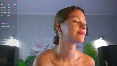 Media: Video of a serene, topless woman with fair skin and light brown hair in a bun, smiling gently. Background features a blurred, minimalist room with a floral painting, green plants, and modern lighting.