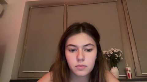 Media: Video of a young Caucasian woman with long brown hair, wearing minimal makeup, sitting indoors. The background features closed beige blinds and a vase with white flowers on a dark countertop.