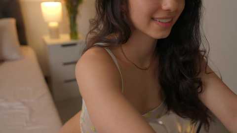 Media: Video of a young woman with long dark hair, wearing a floral bra, smiling, in a softly lit bedroom with a bed and nightstand.