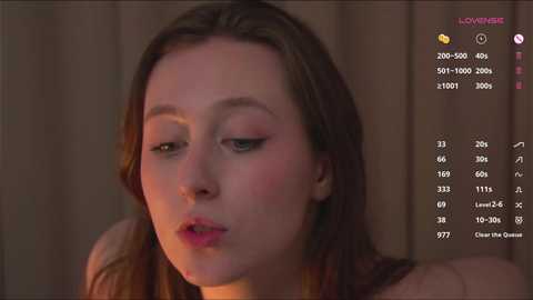 Media: Video of a young Caucasian woman with fair skin, light brown hair, and pink lips, wearing minimal makeup, looking slightly melancholic, with a blurred background.