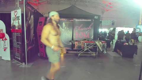 Media: Video of a shirtless, muscular man with a gray cap and green shorts walking past a crowded outdoor event with tents, banners, and people.